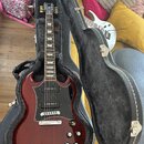 Červená elektrická kytara s černým pickguardem v černém tvrdém kufru s šedou vnitřní výstuhou. Na hlavě hmatníku je bílé logo Gibson. Nástroj má dva…