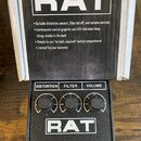 Černý efekt pedál s nápisem RAT a třemi ovládacími knoflíky označenými jako DISTORTION, FILTER a VOLUME. Pedál je umístěn před otevřenou krabicí, na…