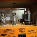 Fotografie zachycuje vnitřek kytarového zesilovače s viditelnými elektronkami a ovládacím panelem. Na oranžovém panelu jsou nápisy WARNING! ALWAYS CO…