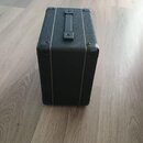 Černý obdélníkový kufr z texturovaného materiálu s plastovou rukojetí. Rohy jsou chráněny kovovými prvky a hrany jsou zakončeny světlými lištami. Na…