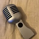 Stříbrný mikrofon v retro stylu s mřížkou z horizontálních slotů. Nástroj je vyroben z plastu s matným povrchem a má integrovaný držák. Na viditelnýc…