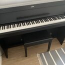 Černý digitální piano s klaviaturou a přívodním kabelem. Na horní části korpusu je bílé logo YAMAHA. Před nástrojem stojí černá pianová stolička. Pov…