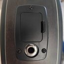 Detailní pohled na černý plastový ovládací panel s bateriovým víčkem a zapuštěným kovovým jack konektorem. Panel je přišroubován třemi křížovými šrou…