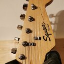 Fotografie zachycuje hlavu elektrické kytary z jasného dřeva s chromovanými ladicími kolíky. Na povrchu je černý nápis Squier by Fender a vedle něj s…