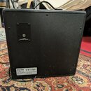 Zadní strana černého kytarového komba s texturovaným povrchem. V horní části je umístěn konektor s nápisem 4 OHM SPEAKER OUTPUT. Dole je přilepený št…