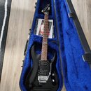 Černá elektrická kytara s hnědým hmatníkem v otevřeném kufru s modrou vnitřní výplní. Na hmatníku je viditelný nápis Ibanez. Kytara má dva humbuckery…