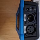 Detail zadní strany modrého elektronického zařízení s černým panelem konektorů. Jsou zde vidět dva XLR konektory s popisky OUT a MIC, USB port a jack…