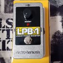 Kytarový efekt v stříbrném kovovém krytu umístěný na žluté krabici. Na zařízení je černý panel s nápisy „LPB-1“, „LINEAR POWER BOOSTER“ a „BOOST“, če…