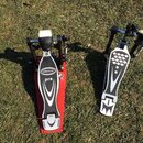 Dvě červeně a stříbrně provedená pedálová zařízení (double bass drum pedals) položená na trávě. Na červeném pedálu je viditelný nápis Millenium a pod…