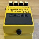 Žlutý efekt pedál s černým horním panelem a čtyřmi bílo-černými ovládacími knoflíky. Na horní části je nápis Over Drive / Distortion OS-2 a logo BOSS…