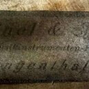 Detailní záběr na kovový štitek s nápisy. V horní části je kurzívou napsáno Meisel & Gerold, pod tím následuje text Musikinstrumenten-Fabrik a v doln…