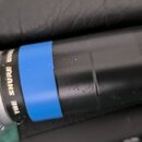 Černý dynamický mikrofon s kovovou stříbrnou mřížkou. Na těle je viditelný nápis THE SHURE WIRELESS a modrý ozdobný pruh. V pravé části jsou tlačítka…