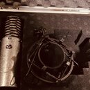 V otevřeném kufru s pěnovou výplní je umístěn stříbrný kondenzátorový mikrofon a černý pavouk (shock mount). Na těle mikrofonu je vidět nápis Spirit…