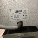 Detail štítku na šedém kovovém zařízení s texturou. Na bílém štítku je logo TRAMAG a nápisy: pri. 220 V, sec. 110 V, L.Nr. D 1601, 50÷60 Hz, 1600 VA,…