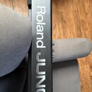 Detailní pohled na černý povrch hudebního nástroje. Je na něm bílý nápis Roland JUNO-DS. Povrch vykazuje drobné škrábance a opotřebení.
