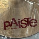 Detailní pohled na mosazný činel v průhledném plastovém obalu. Na povrchu je červeným nápisem uvedena značka pAiste a pod ní text MADE IN SWITZERLAND…