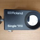 Černý trigger s nápisy Roland a Single TRIGGER RT-30H. Zařízení má obdélníkový tvar s vykrouženým otvorem a je vybaveno šroubovacím knoflíkem. Povrch…