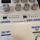 Bílý efekt pedál s nápisy BOSS, DIGITAL DELAY a DD-200. Na horní části jsou umístěny stříbrné ovládací knoflíky a několik malých černých tlačítek u s…