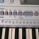 Detailní pohled na ovládací panel a klávesy elektronického klavíru stříbrné barvy. Na panelu jsou viditelné nápisy MASTER VOLUME, STYLE, ACMP, SYNC S…