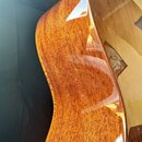 Detailní pohled na bok a zadní část akustické kytary z reddish-brown dřeva s výraznou sletou strukturou. Povrch je pokryt vysokým leskem, v němž jsou…