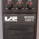 Černý efekt pedál s nápisy VESTA FIRE a DISTORTION DST. Horní panel obsahuje pět otočných potenciometrů s červenými značkami a popisky DISTORTION, OU…