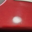 Detailní pohled na červené tělo elektrické kytary s bílým pickguardem a hnedým hmatníkem z javorového dřeva. Povrch laku je lesklý s viditelným odles…