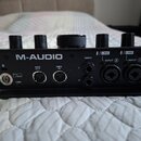 Zadní strana černého audio rozhraní značky M-AUDIO. Jsou zde vidět konektory s popisky MIDI OUT, MIDI IN, MAIN L a R, LINE INPUT 2 a LINE INPUT 1, dá…