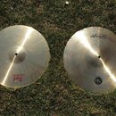 Dvě kovové činely v přirozené zlatavé barvě položené na trávě. Levý činel má nápisy CRASH, CX-200 a logo Pearl. Pravý činel nese nápis Sabian, označe…