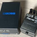 Na fotografii je efekt pedál Reverb značky BOSS, model RV-6, v černé barvě s kovovým tělem. Vedle pedálu je umístěna originální černá krabice s modrý…