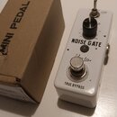 Bílý efekt pedál s nápisem NOISE GATE a logem Rowin. Obsahuje jeden černý ovládací knoflík, jeden přepínač a stomeč s nápisem TRUE BYPASS. Vedle pedá…