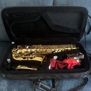 Zlatý saxofon v černém polstrovaném pouzdře. K nástroji je přiložen munčnípíst s korkem, červený látkový potah a krabička s nápisem "Vandoren". Na tě…