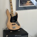 Elektrická basová kytara s přírodním dřevěným povrchem, černým pickguardem a hmatníkem s černými vložkami. Na hlavě nástroje je nápis "Fender Custom…