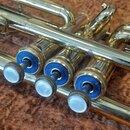 Detailní pohled na mosaznou trumpetu s třemi ventily. Ventily mají modré vložky a bílé perleťové tlačítka. Povrch nástroje vykazuje drobné škrábance…