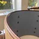 Detailní pohled do vnitřku dřevěného korpusu bubnu. Vnitřní stěna je v šedé barvě a jsou v ní vidět řady kovových šroubů. Horní hrana korpusu vykazuj…