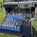 Modrý mixážní pult značky YAMAHA s označením MG10/2 umístěný na černé polici v transportním case. V horní části case je osazen přístroj s nápisy POWE…