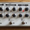 Bílý efektový pedál s čtyřmi sekcemi ovladačů. Na panelu jsou nápisy OVERDRIVE, DISTORTION, DELAY a REVERB, u každé sekce je stisknutý kovový spínač…
