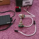 Fotografie zobrazuje sadu elektronických komponentů pro hudební nástroj. Soubor obsahuje černý obdélníkový modul s bílým nápisem EMG, dva potenciomet…