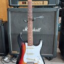 Elektrická kytara s tvarem Stratocaster, v barevné gradaci od žluté přes oranžovou po černou. Na hlavě hmatníku je viditelné logo Fender. Kytara má b…