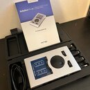 Stříbrné USB audio rozhraní v černém plastovém kufru. Na zařízení je nápis RME Babyface Pro FS a modrý panel s popisky vstupů a výstupů. Vedle přístr…