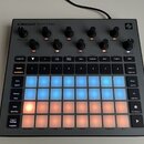 Šedý sampler s nápisem CIRCUIT RHYTHM. Zařízení obsahuje několik černých otočných ovladačů a mřížku podsvícených padů v modré a oranžové barvě. Na po…