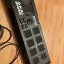 Černý samplovací kontrolér s osmi šedými padovými tlačítky a malým displejem. Na horní části je bílé logo AKAI Professional a nápis MPX8 MOBILE SD SA…