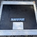 Černý transportní kufr s hliníkovými hranami a rohy. Uprostřed víka je nalepené modrobílé logo SHURE®. Povrch kufrů vykazuje známky opotřebení v podo…