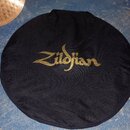 Černý kulatý textilní obal na činely. Uprostřed je zlatým potiskem nápis Zildjian. V horní části jsou vidět dvě úchytná uška. V pozadí jsou vidět čás…