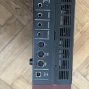 Fotografie zachycuje zadní panel černého elektronického hudebního zařízení. Jsou zde viditelné konektory s popisky OUTPUT (L, BAL, R), PHONES, SUSTAI…