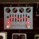 Stříbrný efekt pedál s nápisy ALPHA OMEGA ULTRA a DARKGLASS ELECTRONICS - MADE IN FINLAND. Na horní straně jsou čtyři černé ovládací knoflíky s popis…