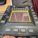 Černý kontrolér KORG Kaossilator Pro+. Uprostřed se nachází čtvercový dotykový panel s žlutým rámečkem. Na přístroji jsou viditelné tlačítka s nápisy…