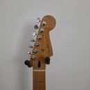 Fotografie zachycuje hlavu a část hmatníku elektrické kytary z přírodního dřeva. Na hlavě je viditelné logo Fender a nápis STRATOCASTER. Nástroj má š…
