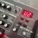 Detailní pohled na ovládací panel efektového pedálu stříbrné barvy. Na panelu je červené digitální displej s číslem 02 a nápisy REVERB, DELAY, OUTPUT…