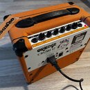 Kytarové kombo v oranžovém vinylu s černou rukojetí a plastovými rohy. Na předním panelu jsou ovládací knoflíky a nápisy INPUT, GAIN, DRIVE, BASS, MI…