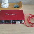 Červené audio rozhraní s nápisem Focusrite na horní straně. Na předním panelu jsou dva vstupy, ovladače zisku a knoflík s nápisy MONITOR a DIRECT MON…